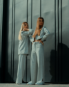Two women in matching light gray tailored suits, standing confidently against a modern, dark vertical panel backdrop